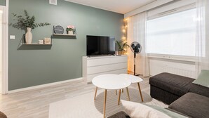 A living room with a flat-screen TV, white storage unit, two white round tables, a dark brown sofa, and a window with blinds.
