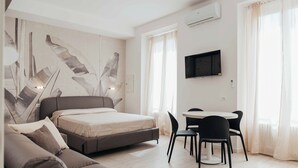 A modern hotel room with a bed, a sofa, a dining table with chairs, a TV, and large windows with curtains.