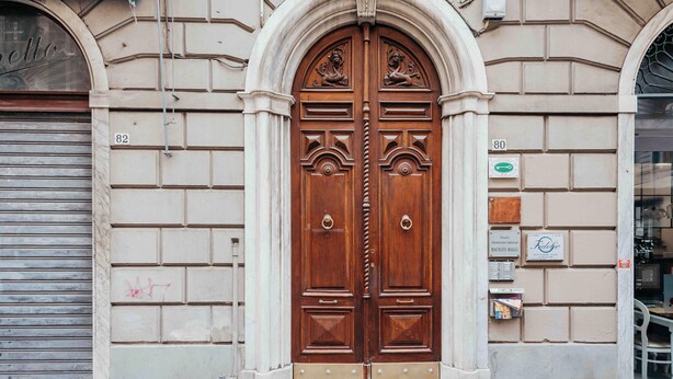 A large wooden double door with intricate carvings, flanked by a stone archway and a metal gate.