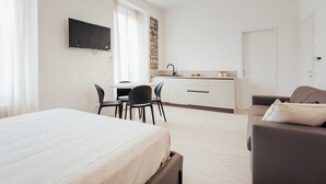 A modern hotel room with a bed, a desk, a chair, a sofa, and a kitchen area.