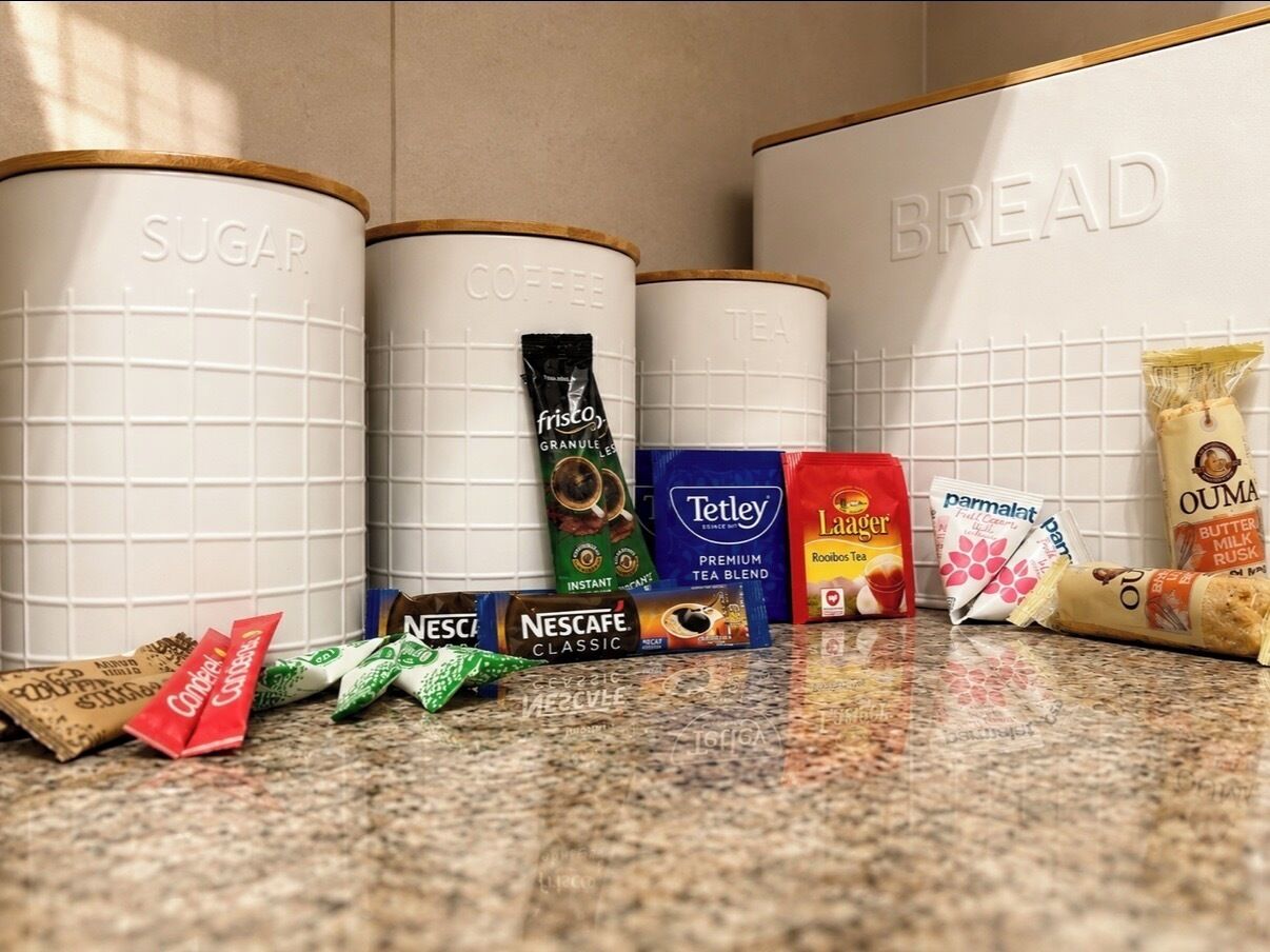 Contenedores de almacenamiento de cocina etiquetados 'SUGAR', 'COFFEE' y 'BREAD' sobre una encimera con diversos alimentos.