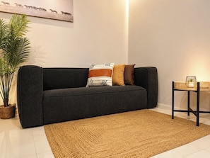 A dark grey sofa with patterned cushions, a woven rug, a small table with a plant, and a wall-mounted artwork.
