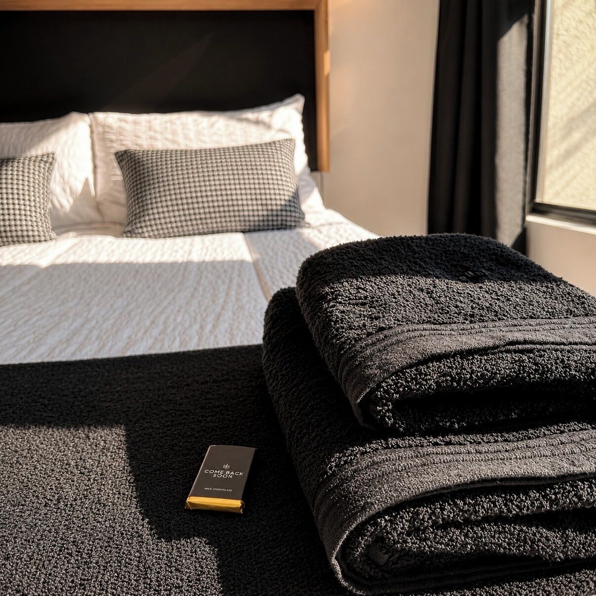 Een net opgemaakt bed met een geruit kussen, een donkere gevouwen handdoek en een chocoladereep op een ribbelruwe vloerkleed.