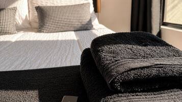 Een net opgemaakt bed met een geruit kussen, een donkere gevouwen handdoek en een chocoladereep op een ribbelruwe vloerkleed.