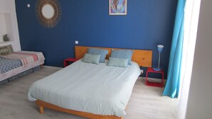 A bedroom with a bed, a nightstand, a red stool, a blue wall, a wooden headboard, a framed picture, and a circular wall decoration. - Gîte in the heart of Suisse Normande  (Saint-Pierre-du-Regard)