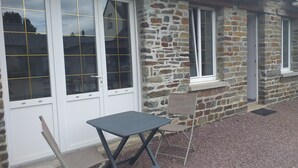 A stone building with white-framed windows and a small table with a chair outside. - Gîte in the heart of Suisse Normande  (Saint-Pierre-du-Regard)