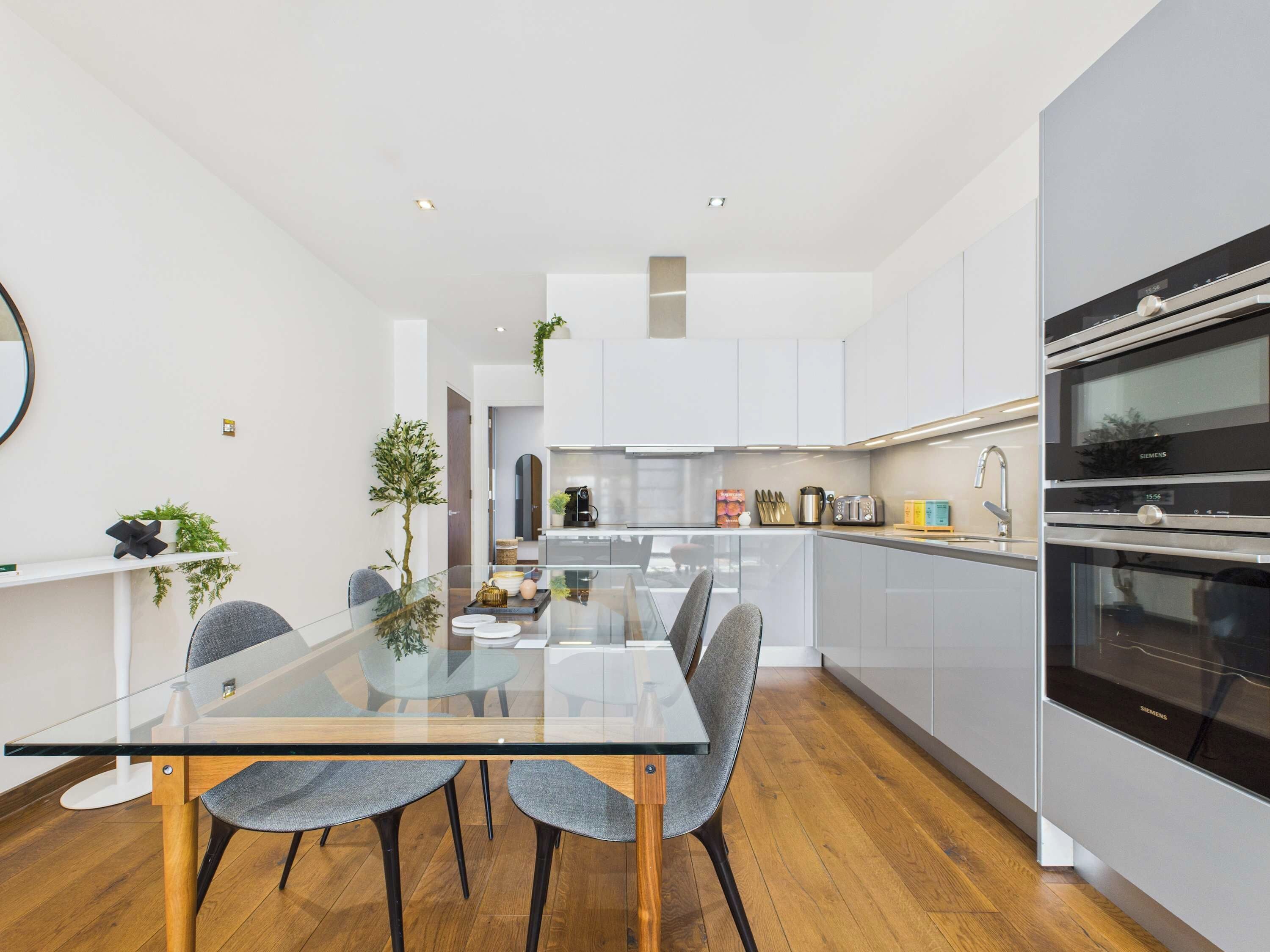 A modern kitchen with white cabinets, a glass dining table, and two grey chairs.