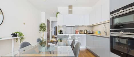 A modern kitchen with white cabinets, a glass dining table, and two grey chairs.