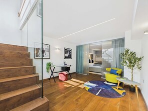 A modern living room with a staircase, a glass partition, a bed, a desk, and a small plant.