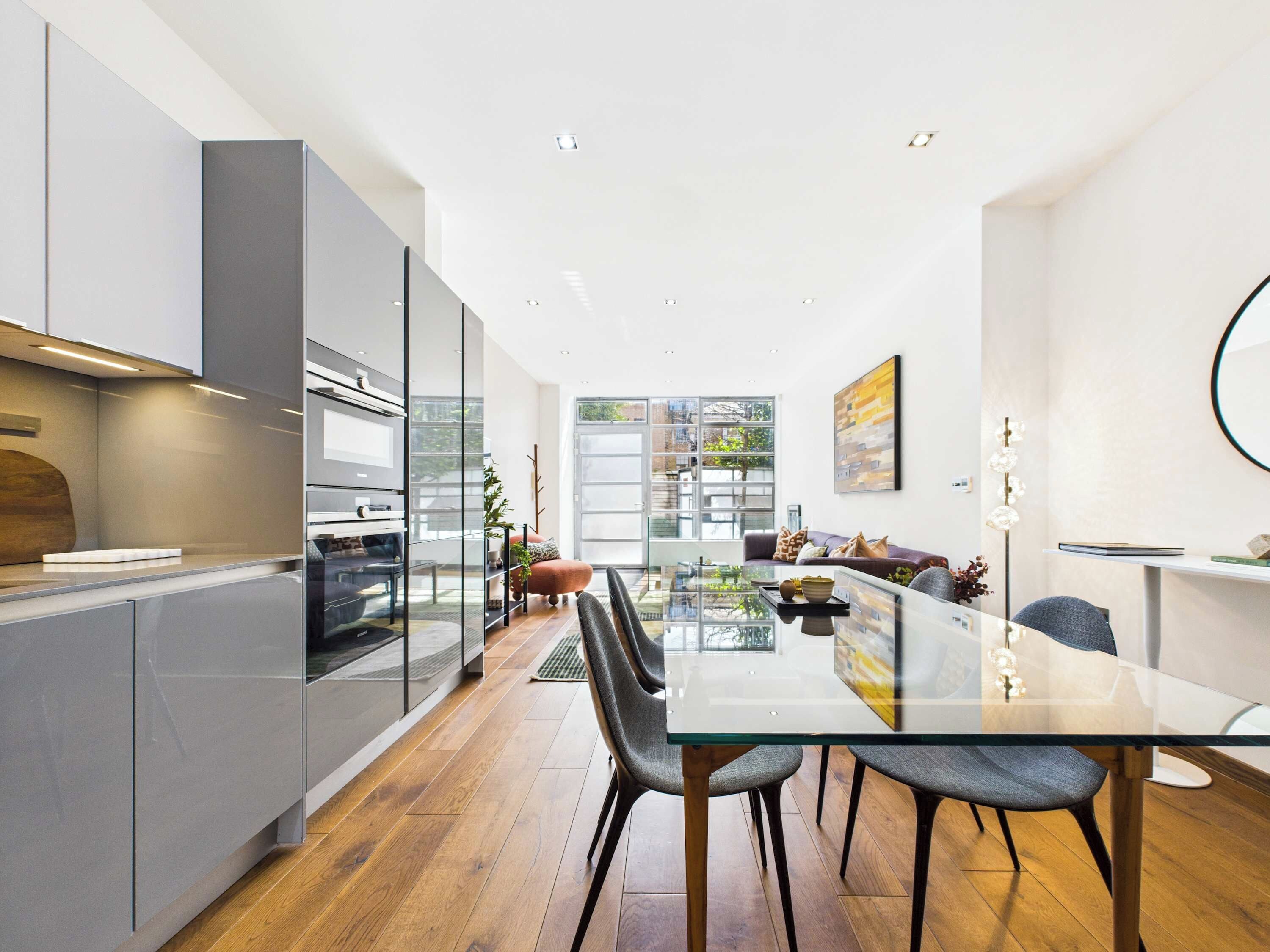 A modern kitchen with a glass dining table, wooden flooring, and a view of the outdoors.