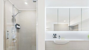 Une salle de bain moderne avec une douche à l’italienne, un lavabo blanc avec un miroir au-dessus, et des toilettes visibles sur la droite.