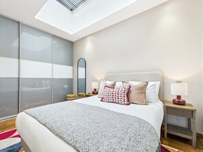 Une chambre à coucher moderne avec une lucarne, un grand lit agrémenté d’oreillers à motifs, des tables de chevet avec des lampes et un grand miroir.