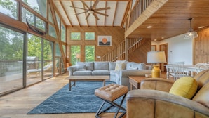 A spacious living area with a large sofa, a coffee table, and a dining table. There is a staircase leading to an upper level, and large windows provide natural light and views of the outdoors. - Kitzbuhl Nook by Innsbrook Vacations! (Innsbrook)