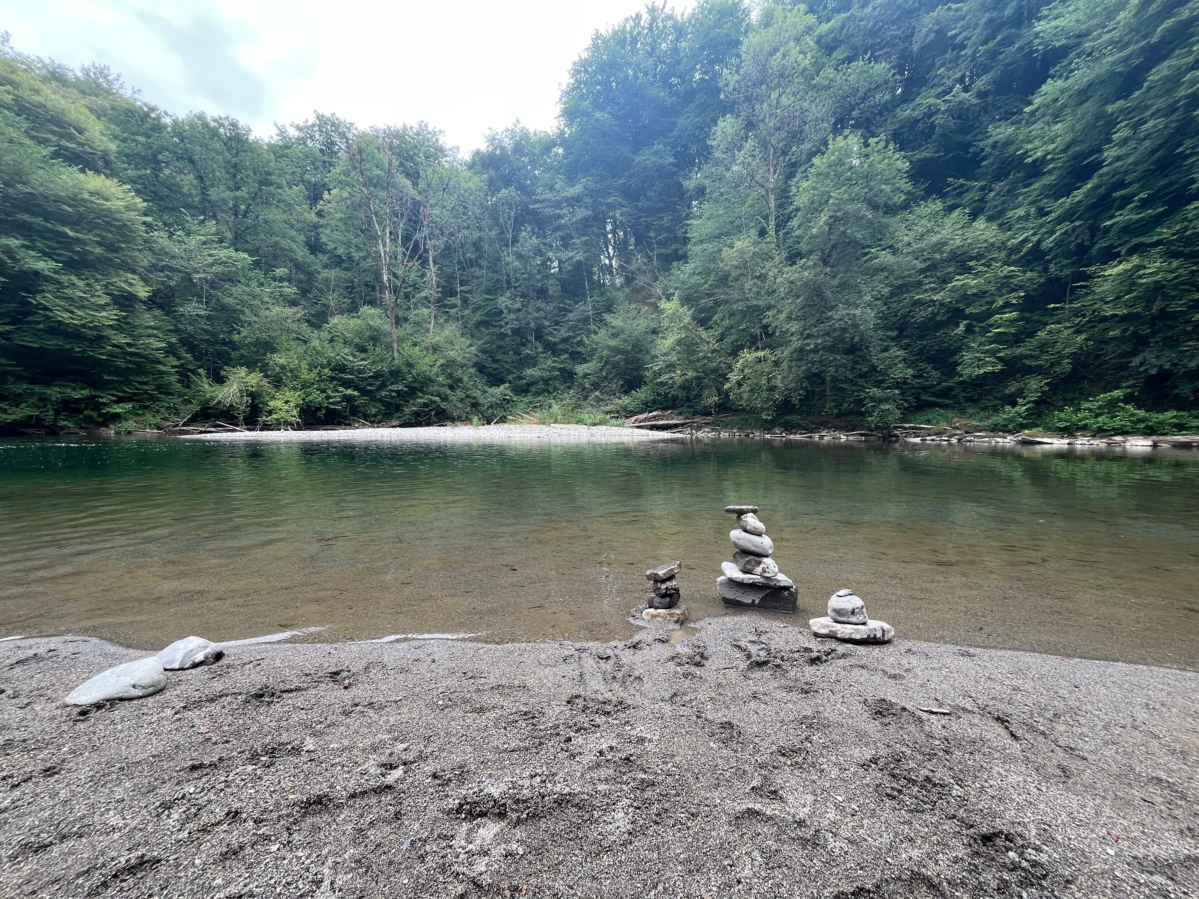 A river surrounded by dense forest, a gravel area with rocks, and a stack of stones.