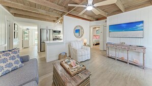 A spacious living area with a high ceiling, a ceiling fan, a large window with a view, a sofa, a coffee table, and a dining area with a table and chairs.
