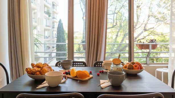A dining table with breakfast items, including croissants, fruits, and coffee cups, set against a backdrop of a balcony with a view of buildings and trees. - L'Ovalino: Quiet, 100m² T4 apartment (Montpellier)