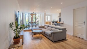 A modern living room with a grey sofa, a round coffee table, and a potted plant. There is a dining area with a table and chairs visible through a glass door. - L'Ovalino: Quiet, 100m² T4 apartment (Montpellier)