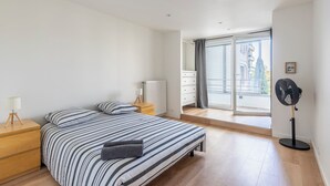 A bedroom with a bed, a bedside table with a lamp, a standing fan, and a view of a building through a large window. - L'Ovalino: Quiet, 100m² T4 apartment (Montpellier)