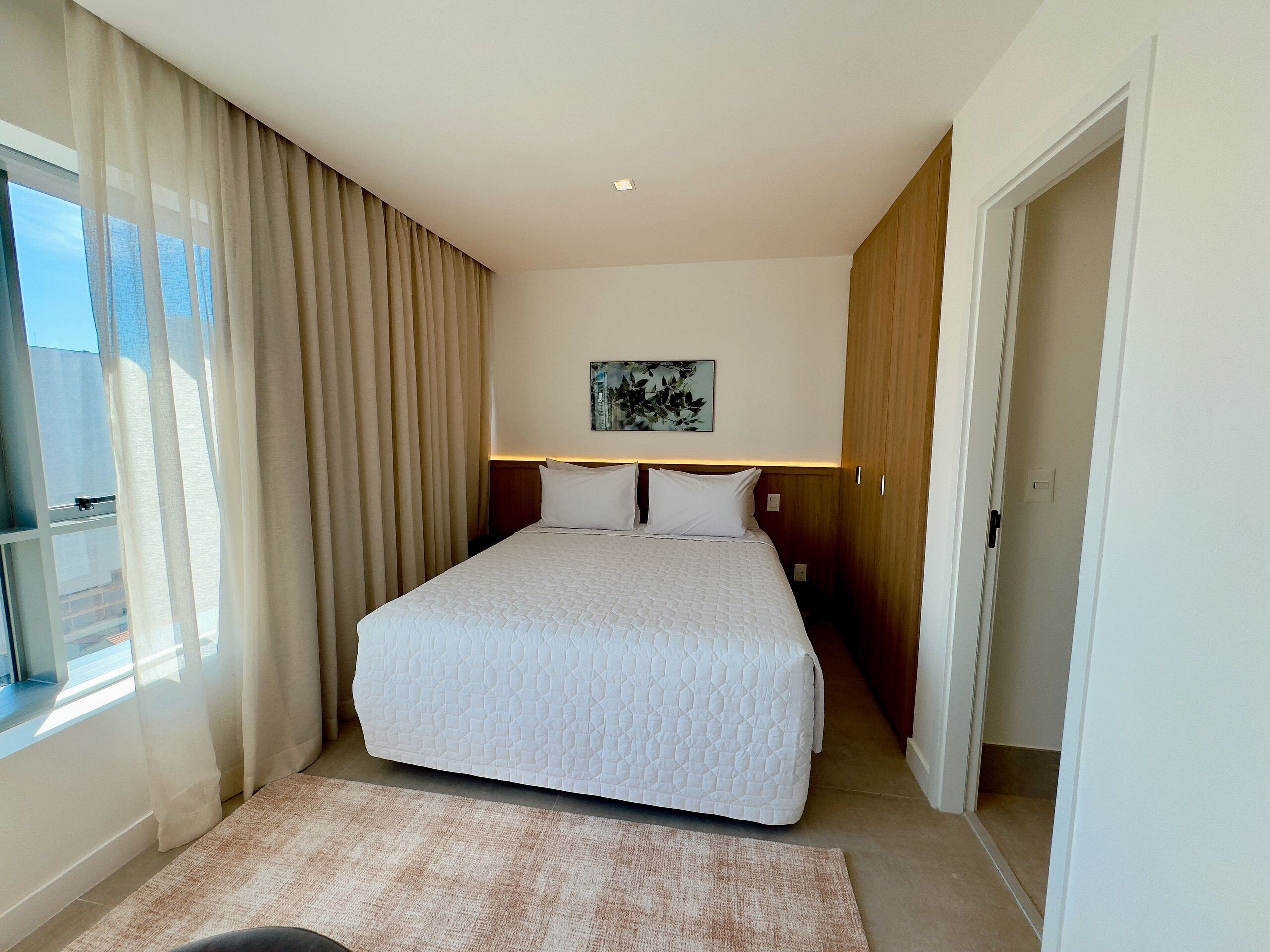 A hotel room with a large bed, a window with curtains, a painting on the wall, and a door.