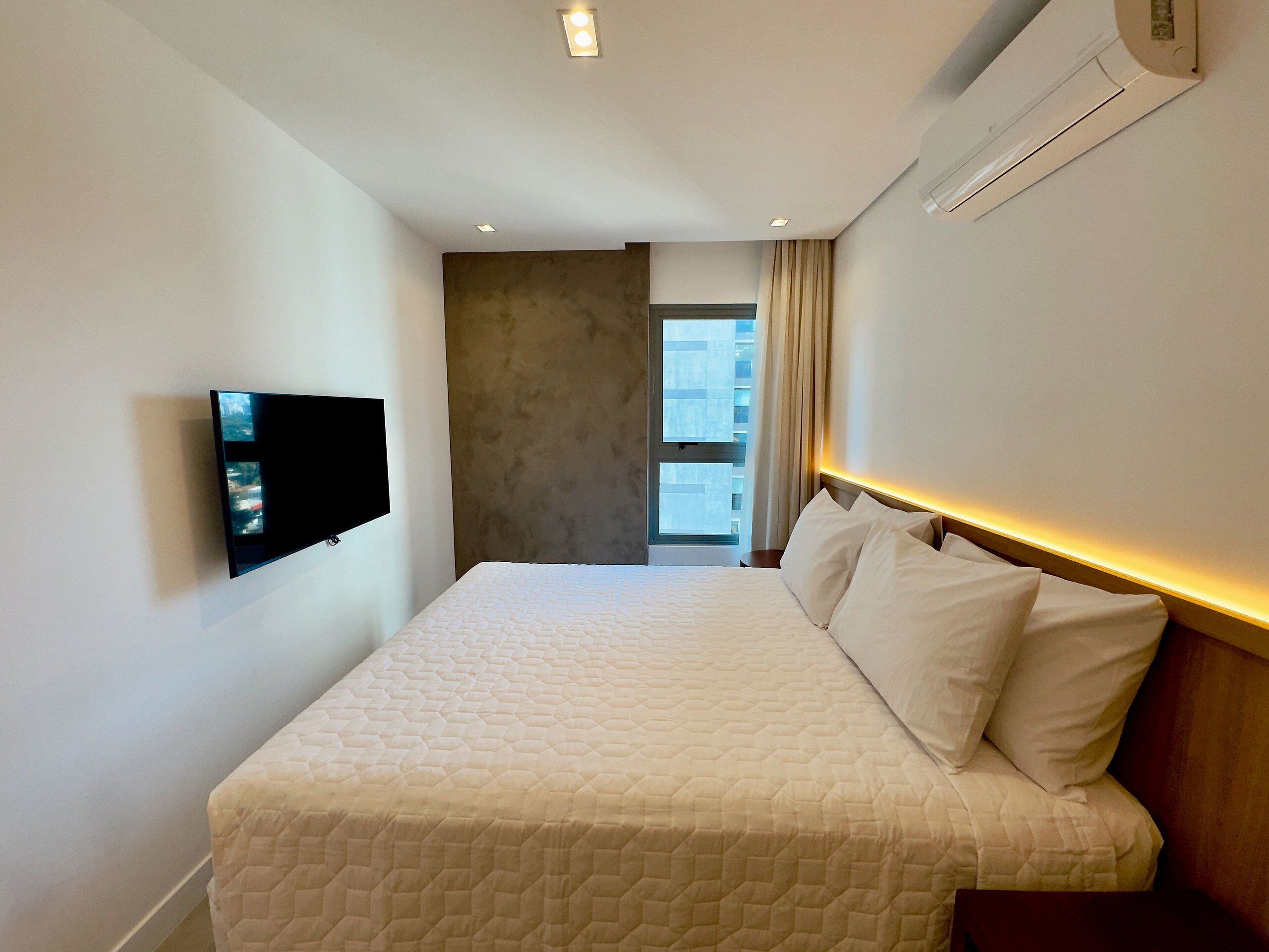 A hotel room with a bed, a flat-screen TV mounted on the wall, and an air conditioning unit.