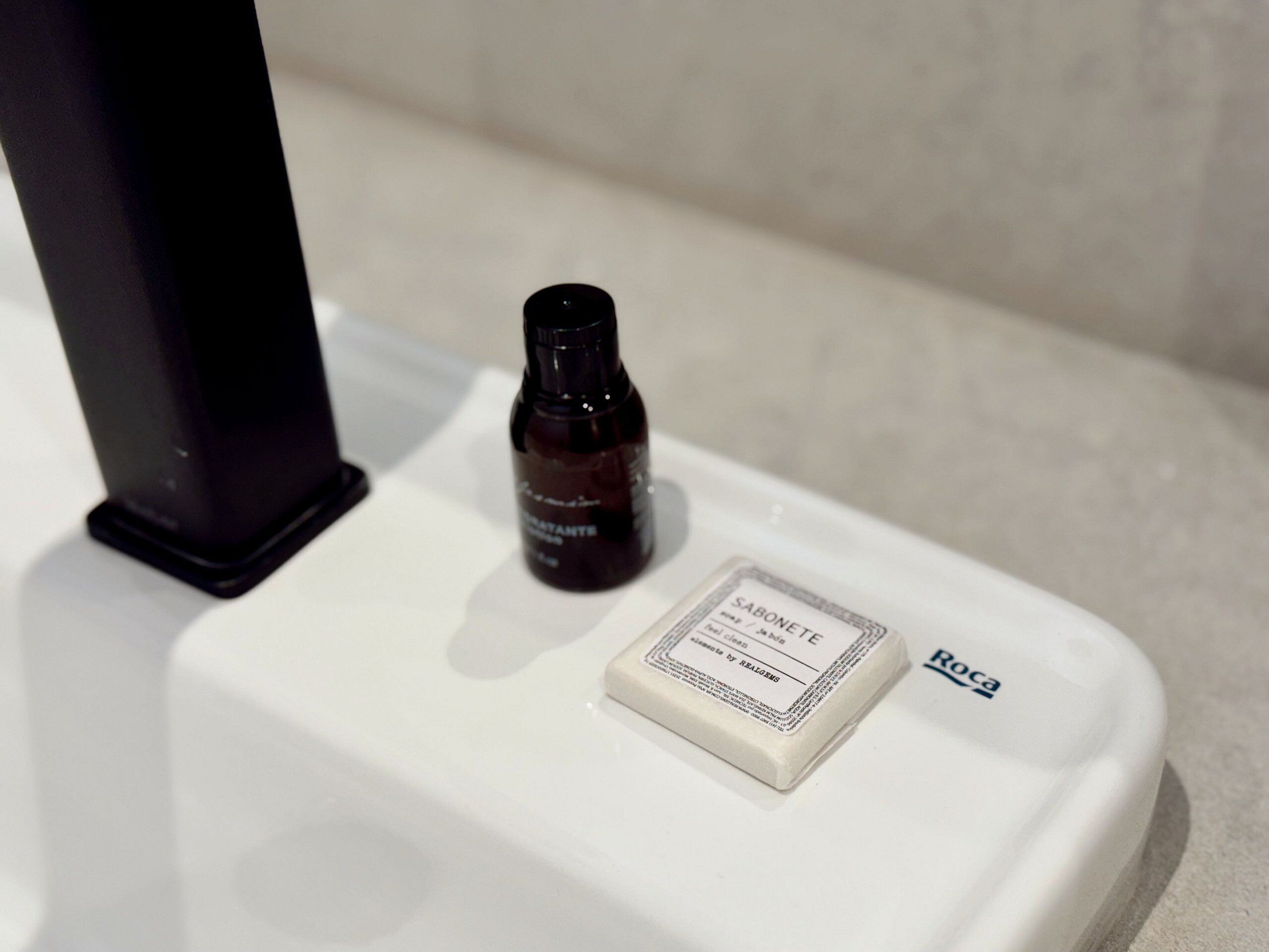 A white bathroom with a black soap dispenser and a soap bar labeled "SABONETE".