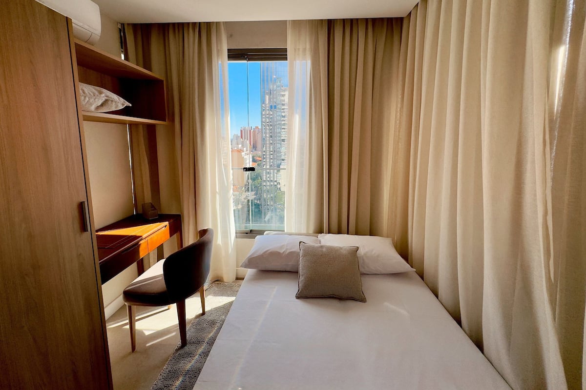 Une chambre d’hôtel équipée d’un lit, d’un bureau, d’une chaise et offrant une vue sur le paysage urbain grâce à de grandes fenêtres.
