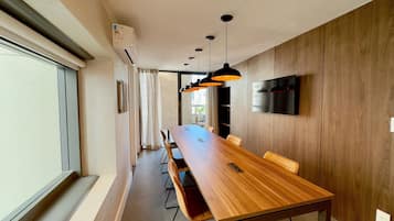 A modern dining area with a long wooden table, black pendant lights, and a flat-screen TV mounted on the wall.