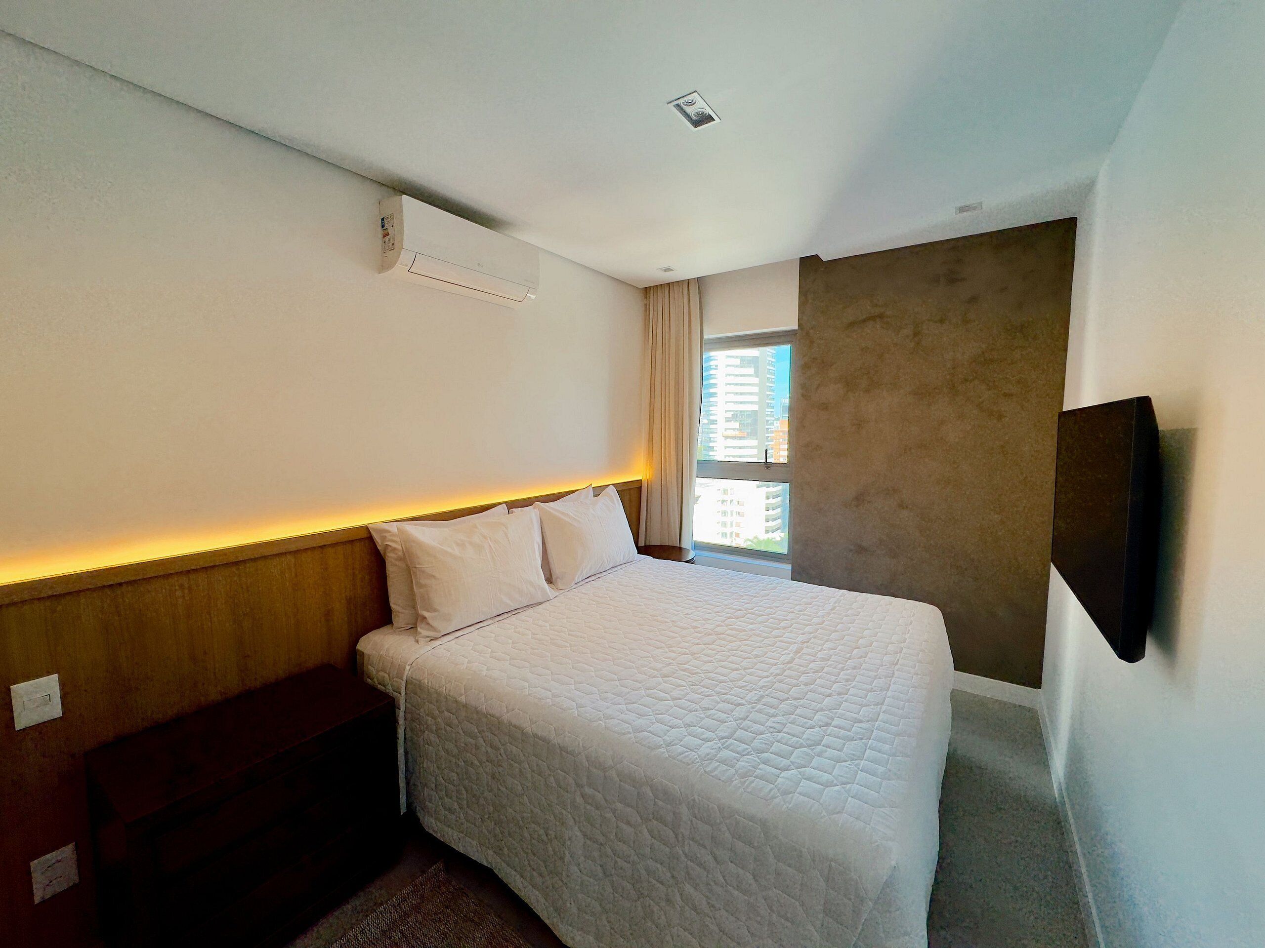 Ein Hotelzimmer mit einem Bett, einem Kopfteil, einem an der Wand montierten Fernseher, einem Fenster mit Vorhängen und einer Deckenleuchte.