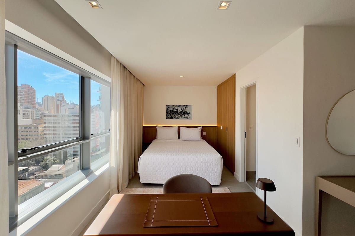 Une chambre d’hôtel avec une grande fenêtre, un lit, un bureau et une chaise.