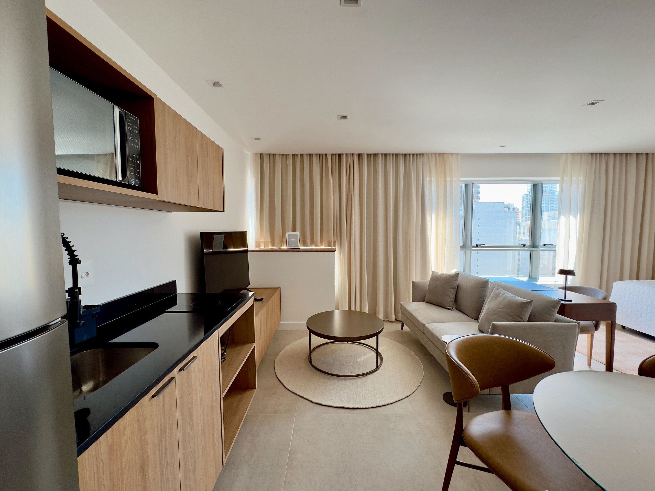 A modern hotel room with a kitchenette, a sofa set, a dining table, and a large window offering a city view.
