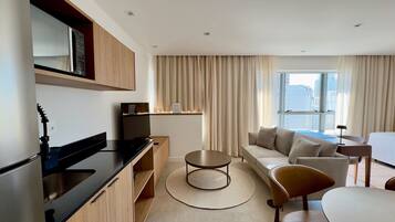 Una habitación de hotel moderna con kitchenette, un conjunto de sofás, una mesa de comedor y un ventanal con vistas a la ciudad.