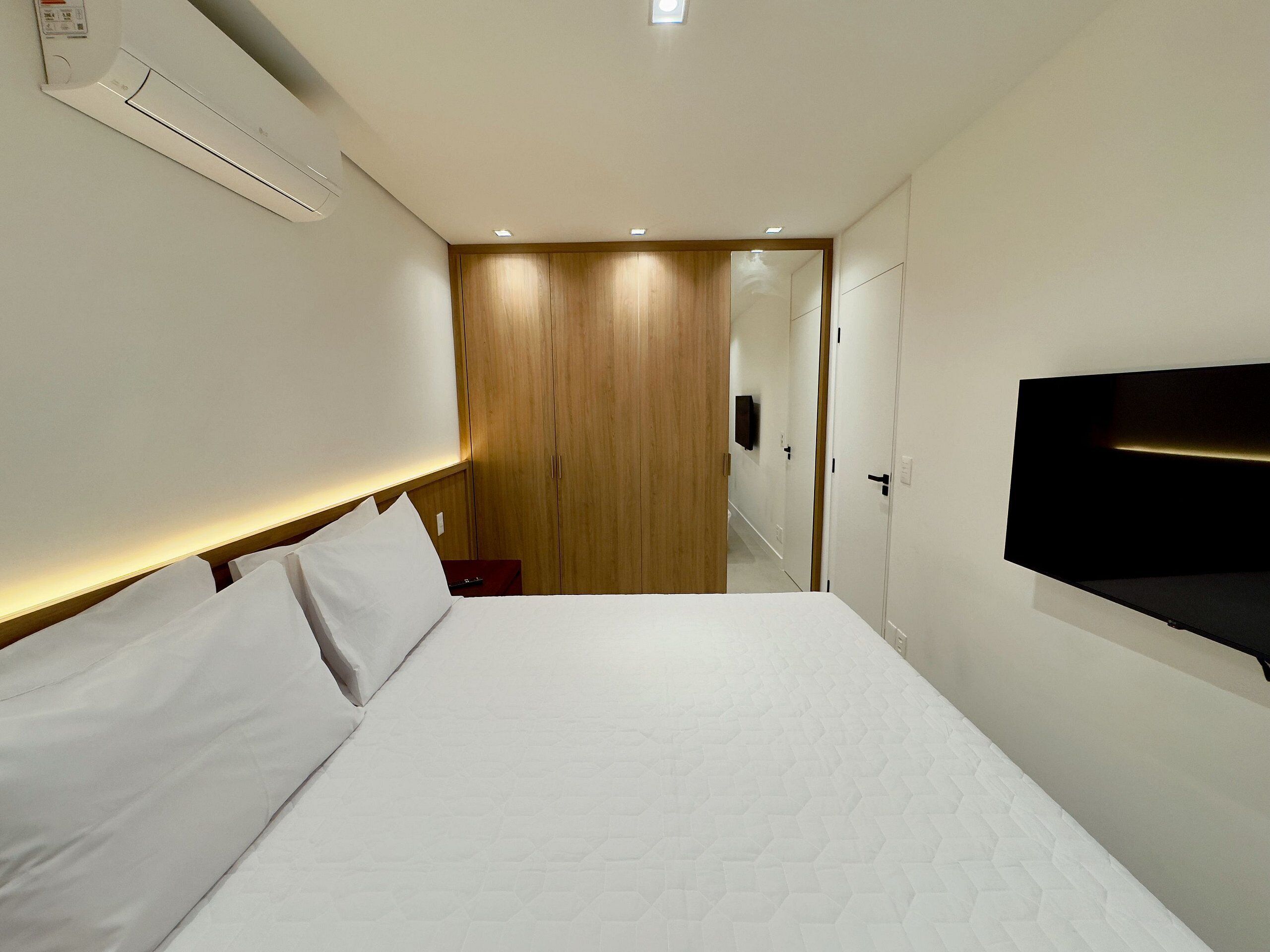 Habitación de hotel con una cama grande, un televisor de pantalla plana en la pared, un armario de madera y una mesita de noche pequeña con un control remoto.