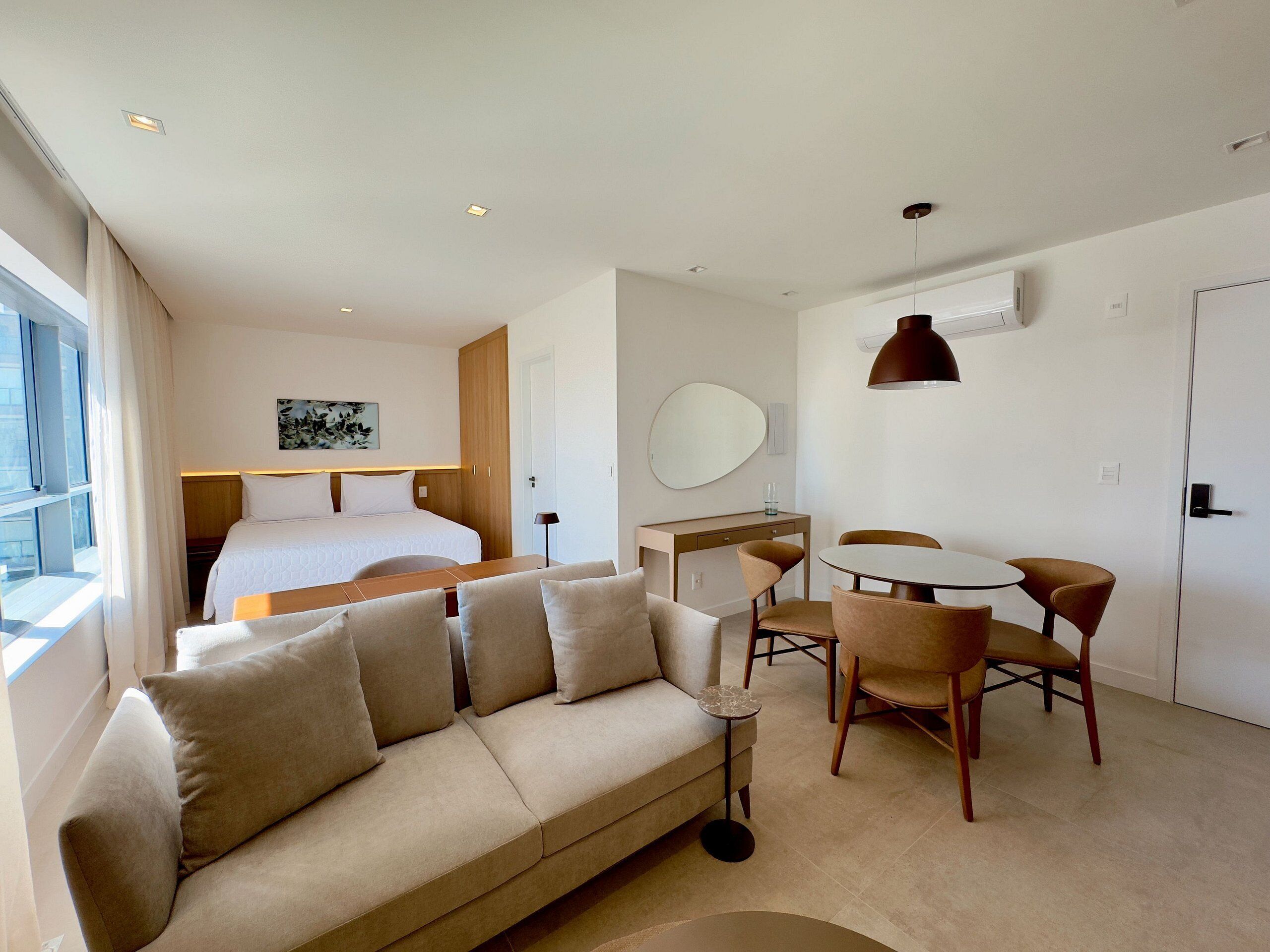 A modern hotel room with a bed, a sofa, a dining table with chairs, a desk, and a large window.
