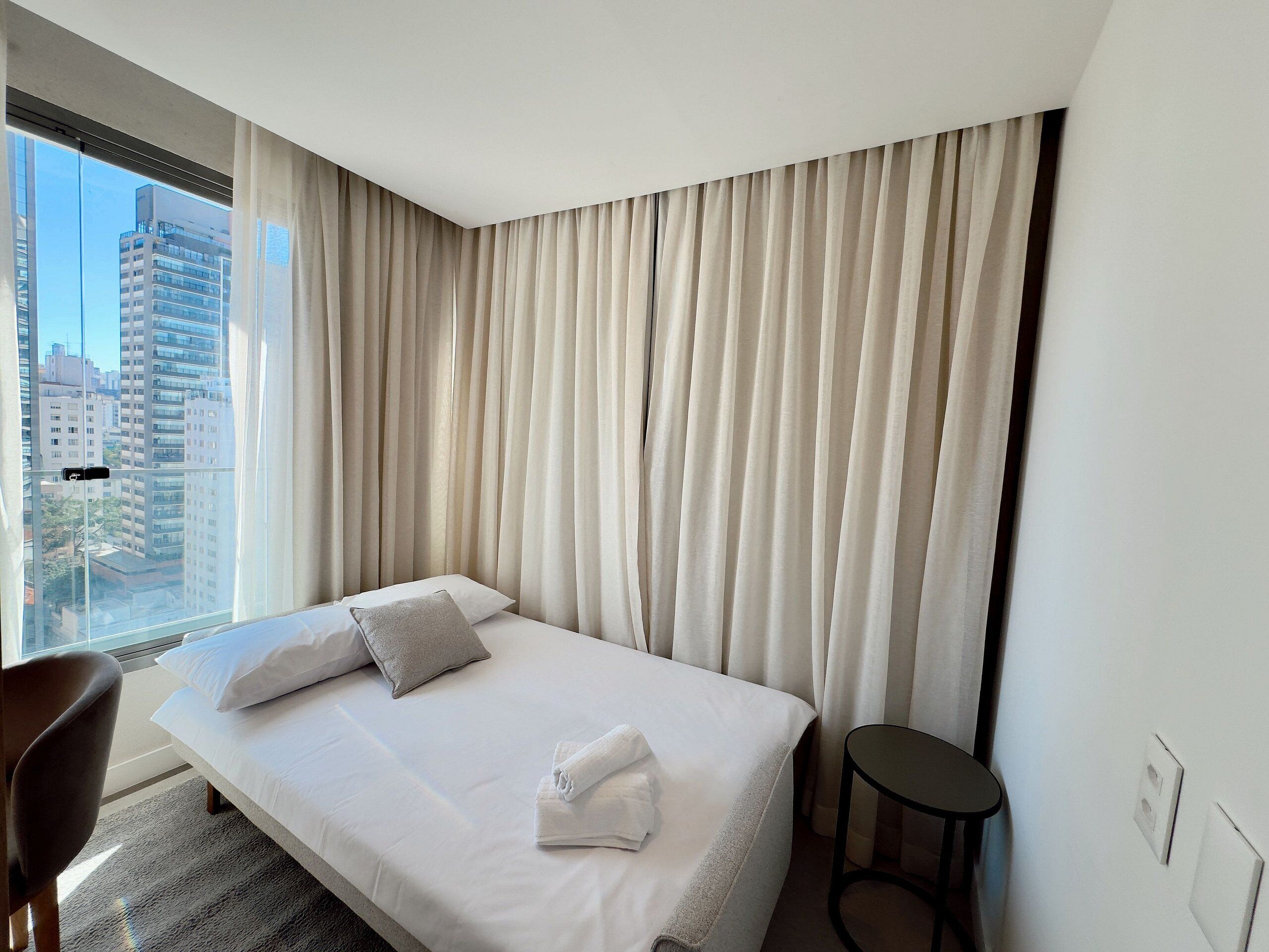 A hotel room with a large bed, a chair, a small table, and a view of the city through the window.