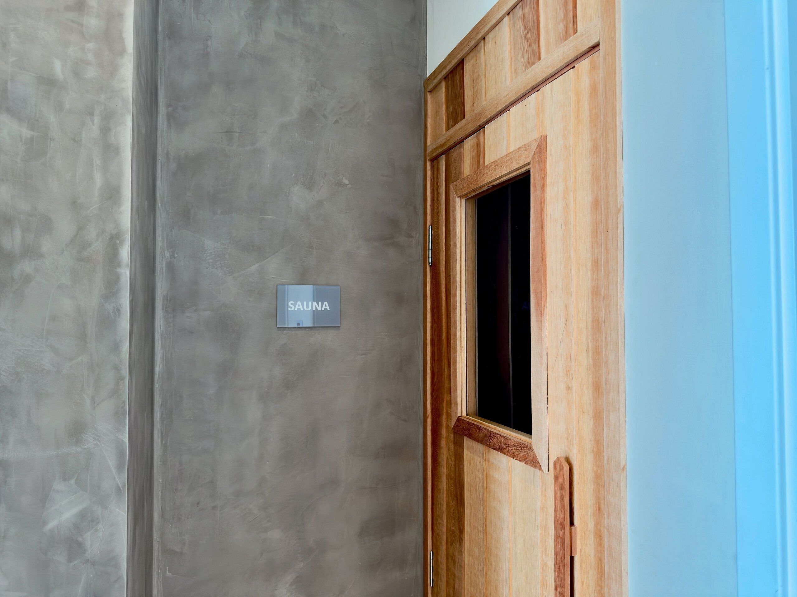 Een houten deur met een bordje waarop ‘SAUNA’ staat, tegen een grijze muur.