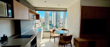 A modern kitchen with a dining area, a large window with a city view, and a flat-screen TV.