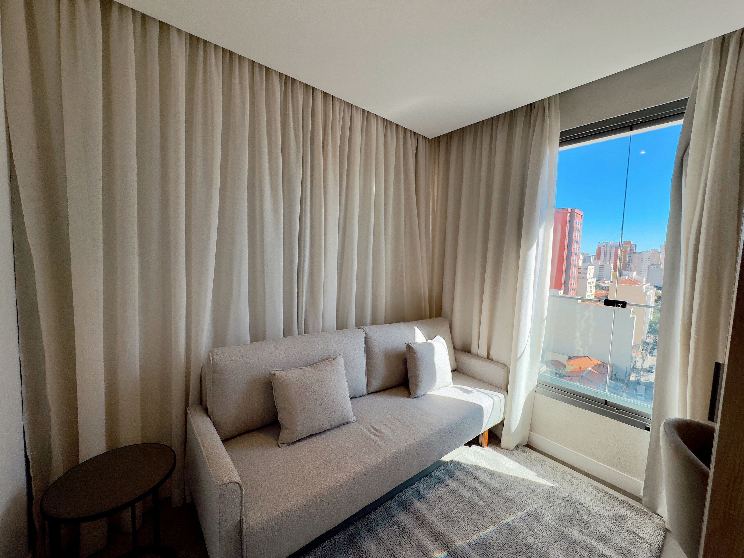 A modern living room with a sofa, a small table, and a view of a cityscape through large windows.