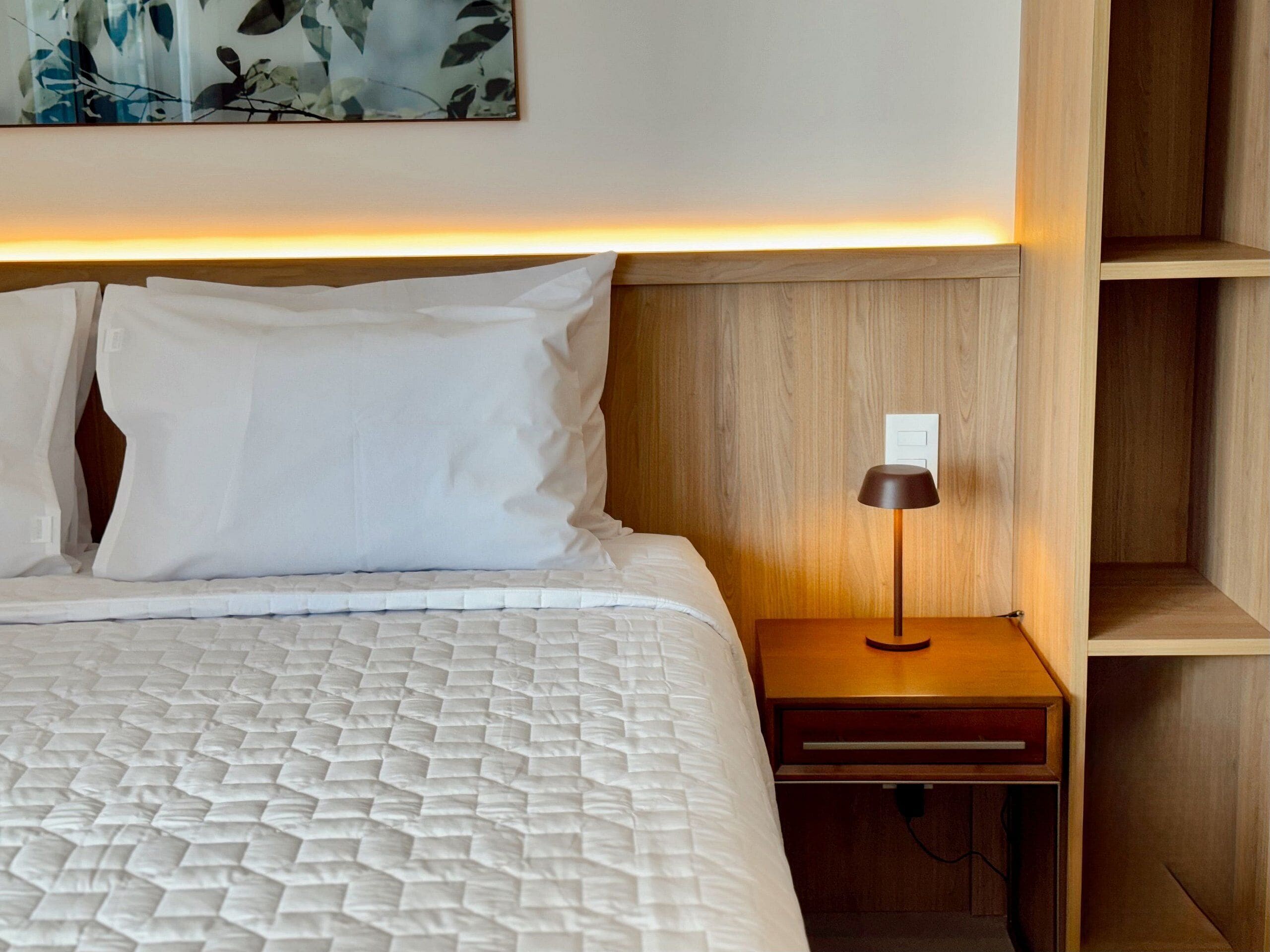 A neatly made bed with white bedding, a bedside lamp, and a wooden nightstand.
