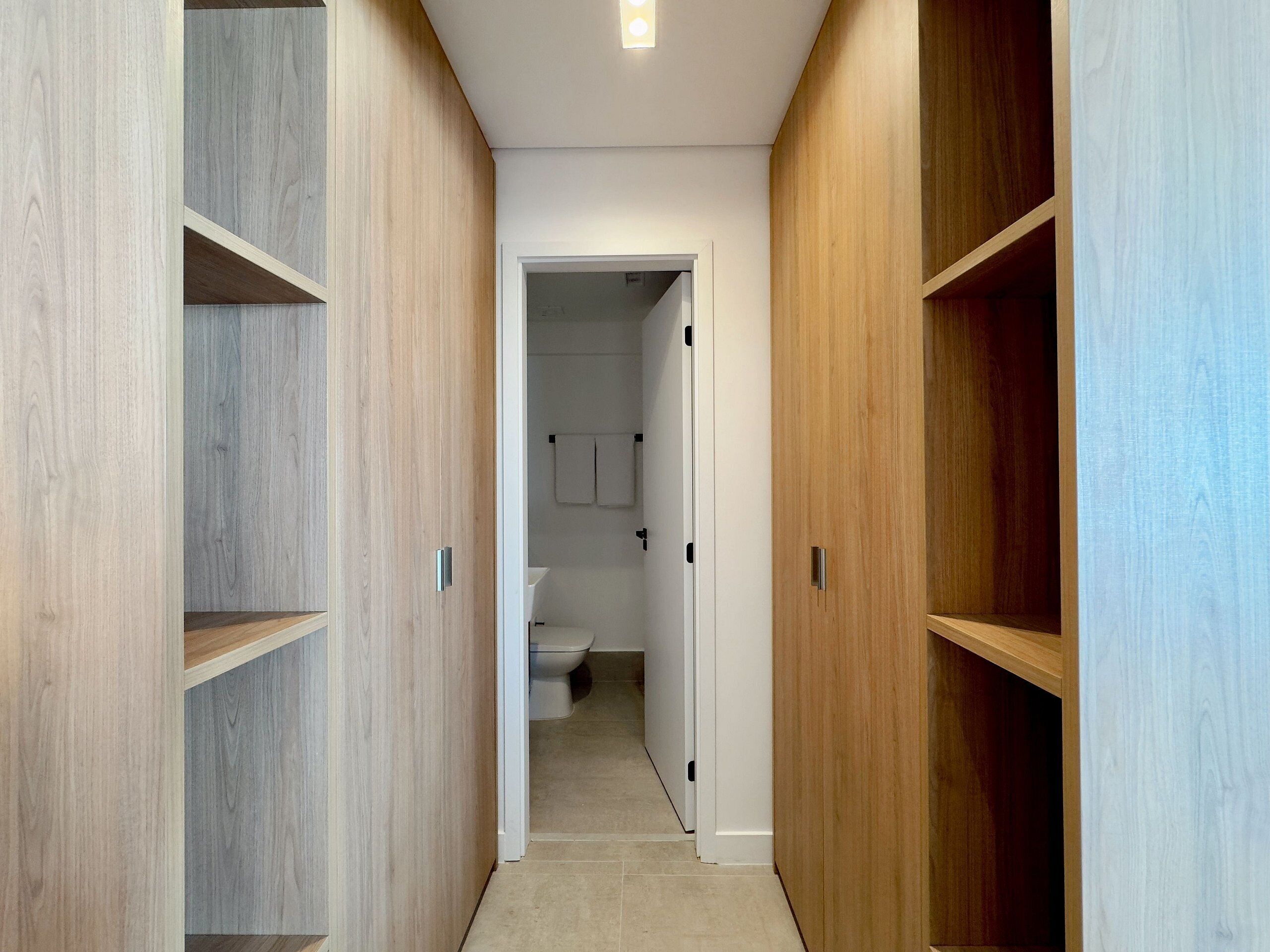 Een smalle gang met houten schapenkasten aan weerszijden, leidend naar een badkamer met toilet en handdoekrek.
