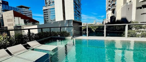 A rooftop pool with clear blue water, surrounded by modern buildings and a glass balustrade.