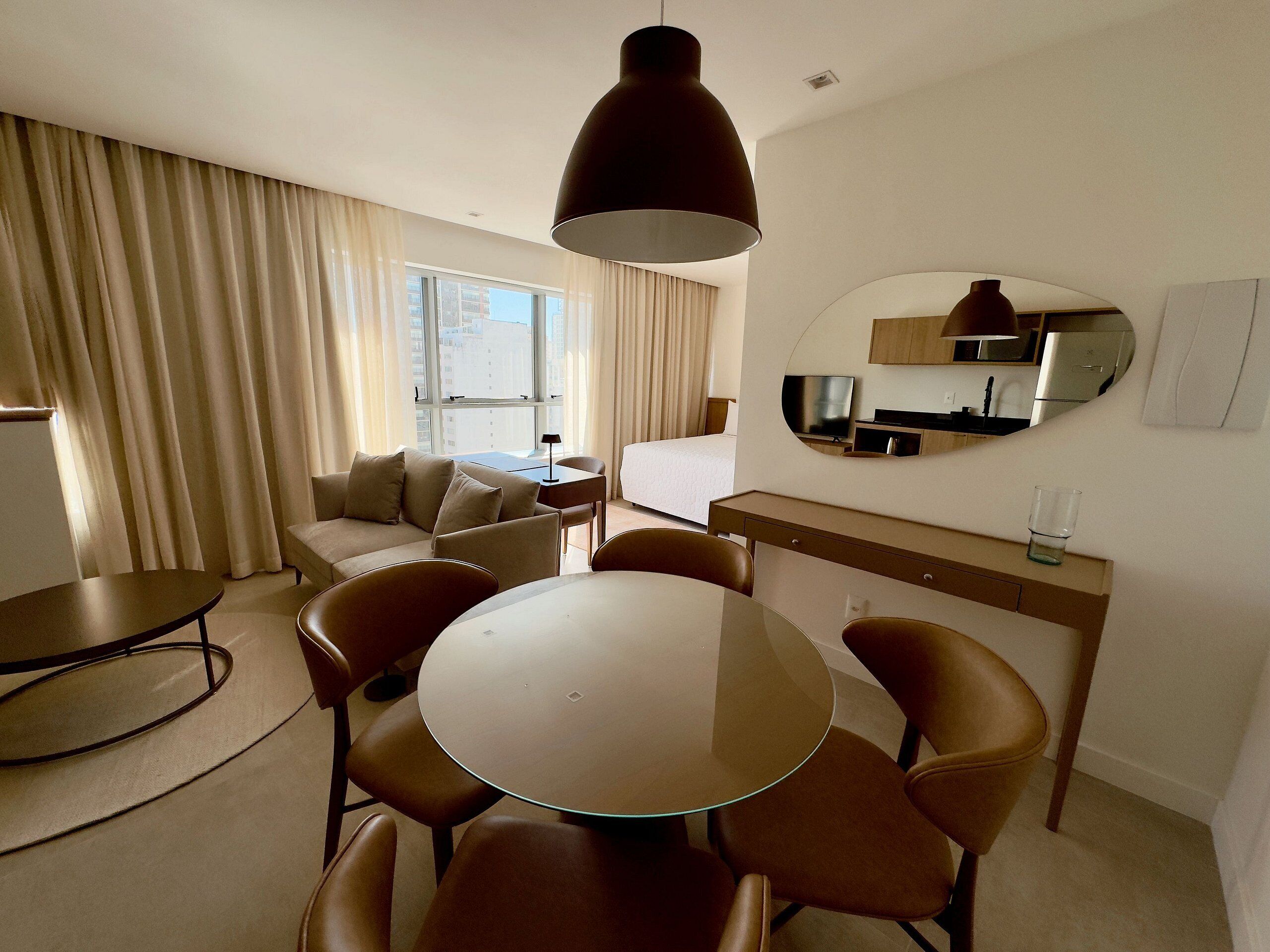A hotel room with a round dining table, six chairs, a sofa, a TV, a minibar, and a large window with curtains.