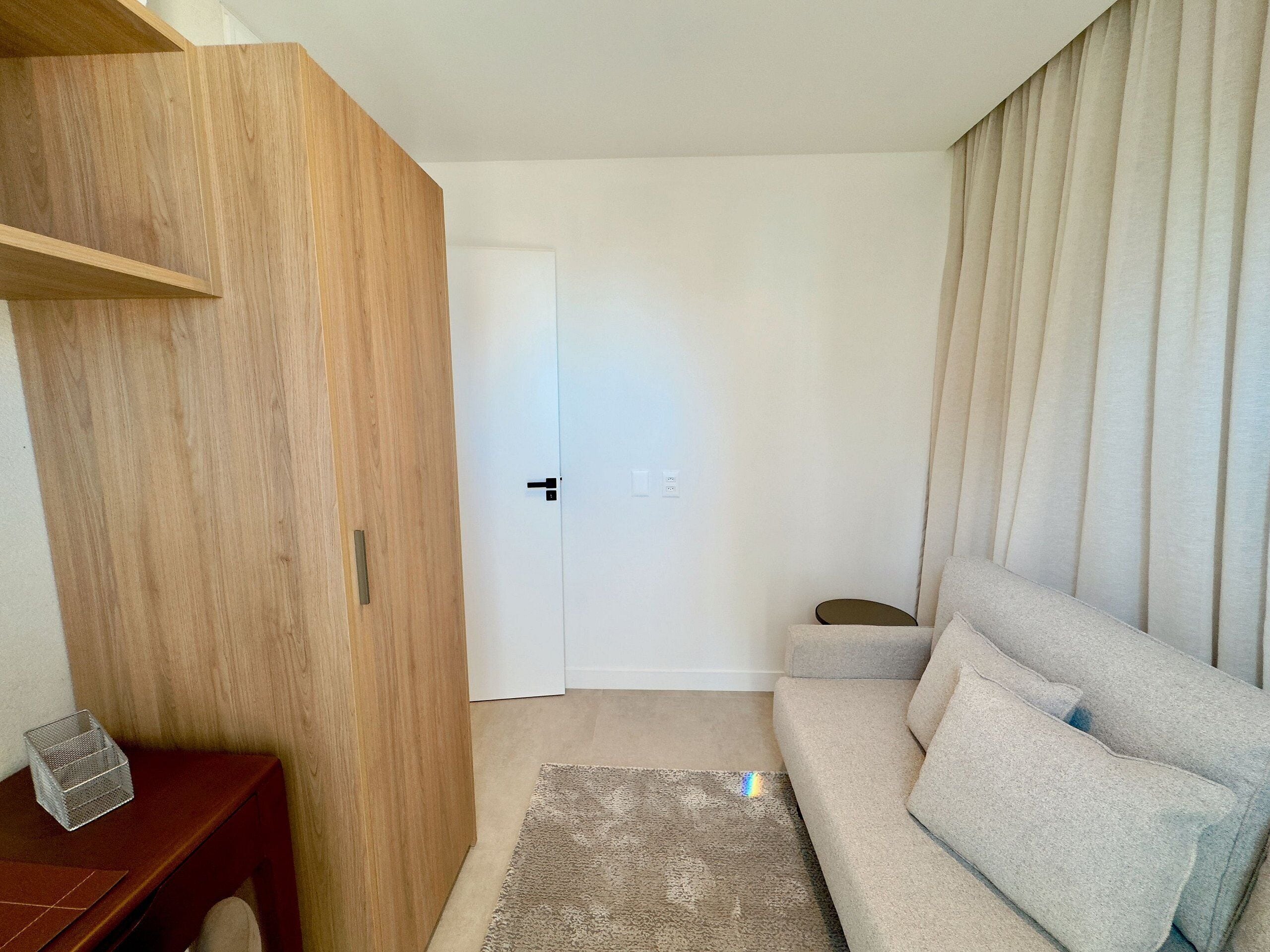 Un espacio habitable pequeño con un armario de madera, un sofá gris, una mesita y una puerta que da a otra habitación.