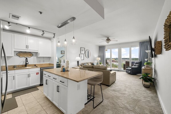 A modern kitchen with white cabinets, a central island, and pendant lighting. There is a dining area with a couch and chairs, and a large window offering a view of the outdoors. - Waterfront resort condo w/ pool and hot tub (Panama City Beach)