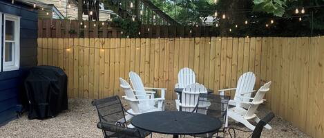 Un patio trasero con una mesa redonda y cuatro sillas Adirondack, rodeado por una valla de madera y luces colgantes.