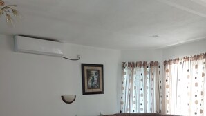 A living room with a brown sectional sofa, a wooden coffee table, a wall-mounted air conditioner, a framed picture, and a window with patterned curtains.