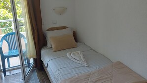 A single bed with a beige quilt, a wall-mounted air conditioner, a small painting, and a blue chair outside the room.