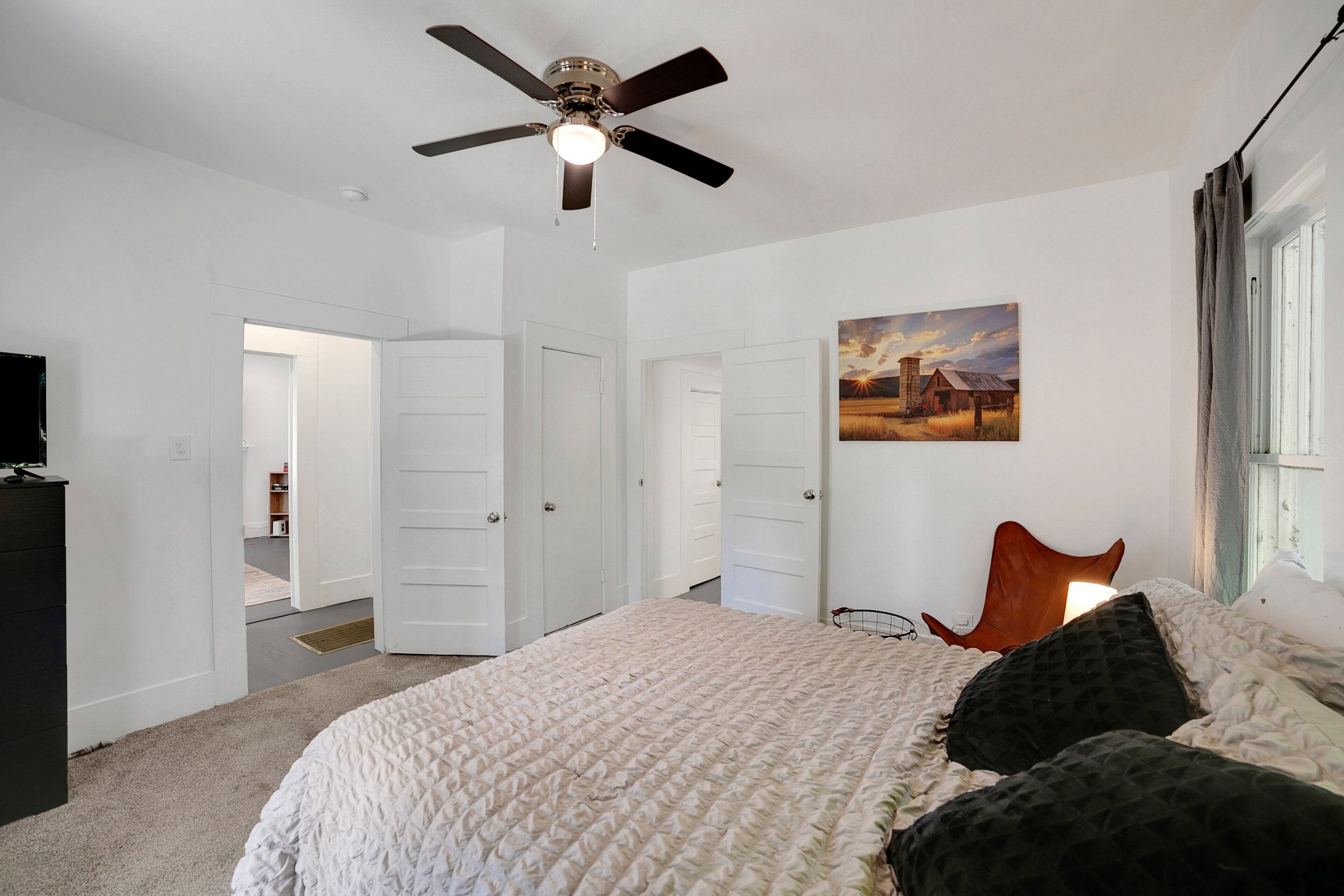 A bedroom with a bed, a ceiling fan, white walls, and a painting.