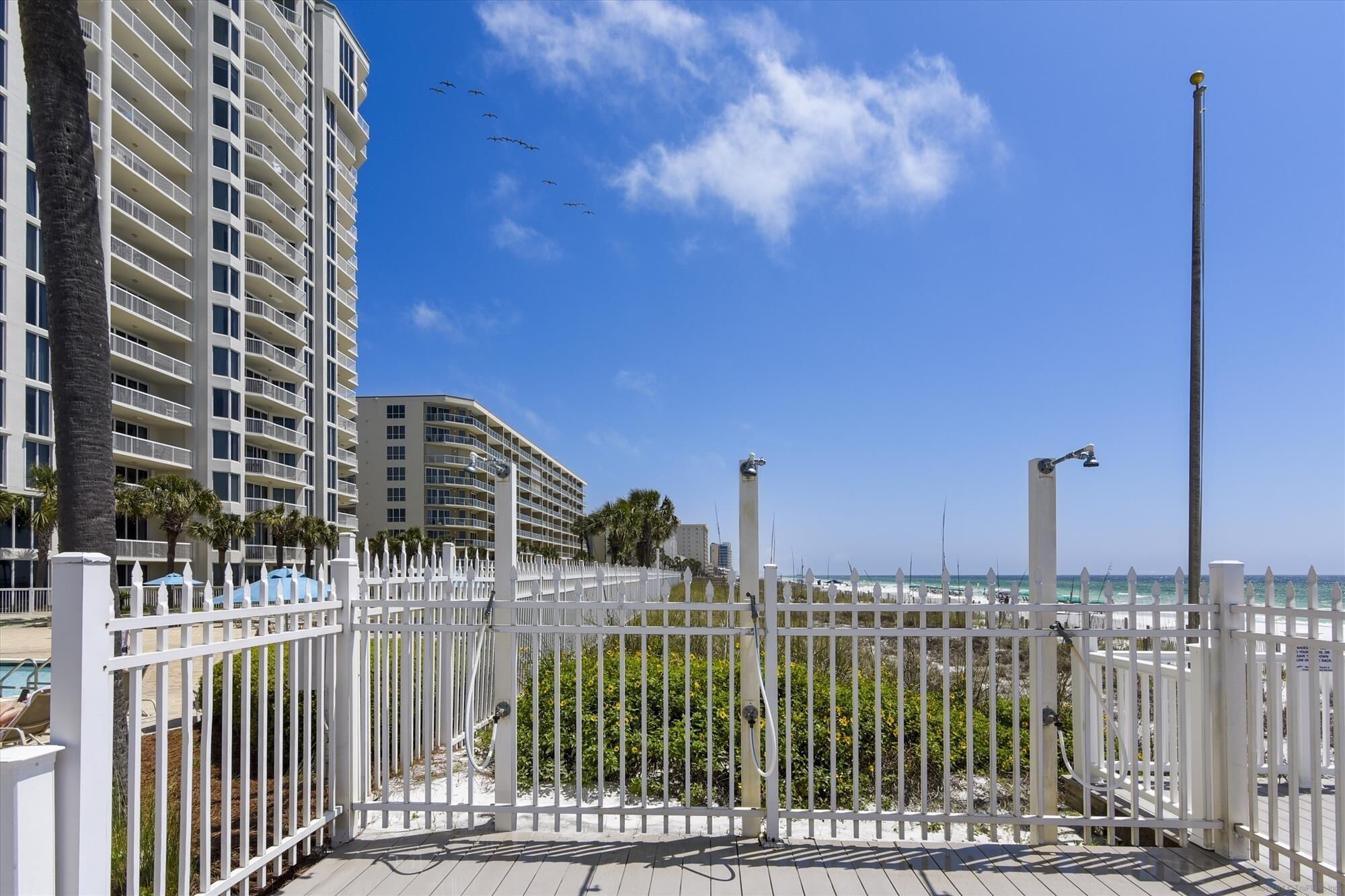A beachfront view with a white fence, a pool area, and high-rise buildings in the background.