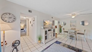 A well-lit, modern living room with a dining area, a large clock, and a TV on a cabinet. - White Sands On Siesta 1 Bedroom Condo (Siesta Key)