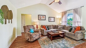 A living room with a ceiling fan, a brown leather armchair, a grey sofa set, a wooden coffee table, and a patterned rug. - 17007 Firenzo Ave 3 Bedroom Home (Panama City Beach)